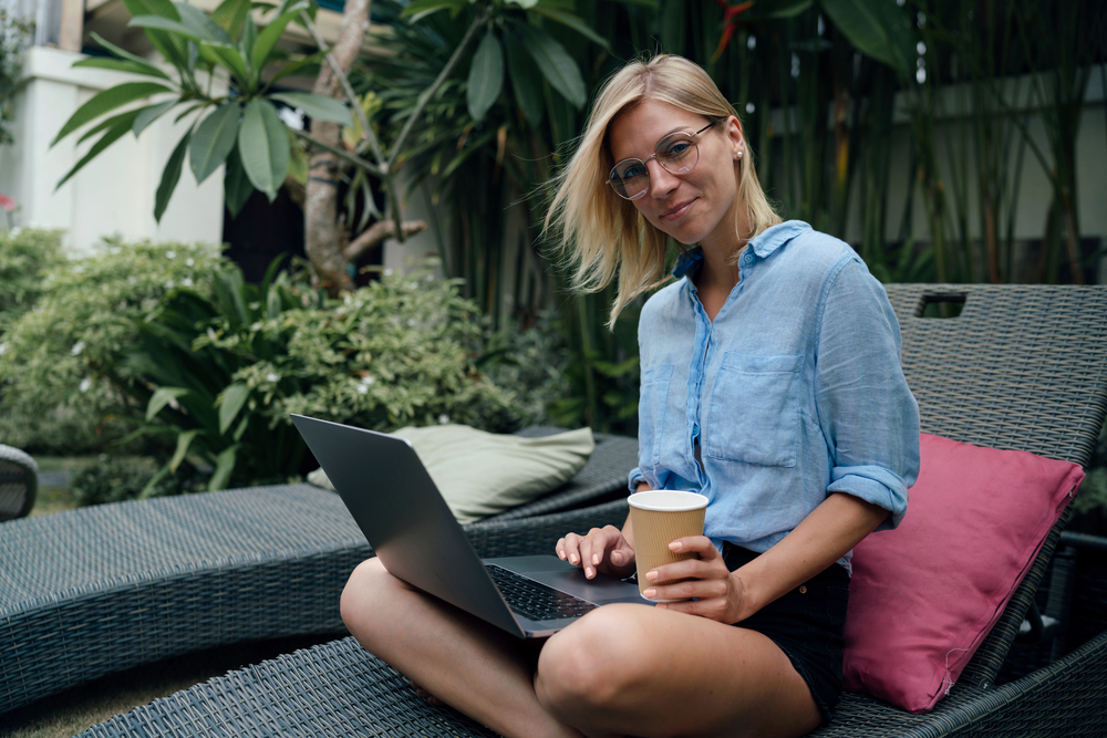 woman with coffee working on a laptop sitting on a lounge chair outside