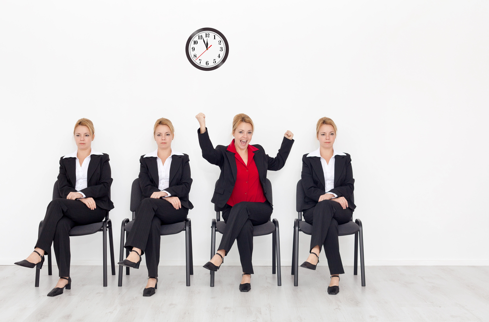 4 women sitting in a waiting room, on dressed in a red shirt and black suit stands out as the others are dressed in white shirts and black suit.