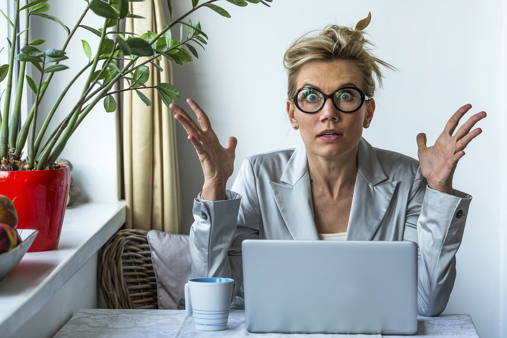Wide eyed woman with her hands in the air as she sits at a laptop trying to write content for her business.